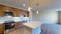 Located at 515 Lincoln Ave in San Jose, California, this single-floor residence showcases a clean, modern design with an open and functional layout. The kitchen is a standout feature, outfitted with warm walnut-toned flat-panel cabinetry extending to the ceiling for generous storage, a crisp white tile backsplash, and light quartz countertops. A central island provides additional workspace and seating, illuminated by elegant pendant lights that complement the recessed ceiling lighting throughout. Stainless steel appliances — including a French door refrigerator, gas range, microwave, and dishwasher — complete the well-appointed kitchen. Wood-look plank flooring flows throughout the main living and kitchen areas, adding warmth and continuity to the space. The bathroom features a full soaking tub and shower combination, a wood-accented vanity with a white countertop, a large framed mirror, and matching wood-look flooring. The bedroom is carpeted for comfort and benefits from abundant natural light through large windows and a glass door that opens directly to a private outdoor balcony, offering views of the surrounding residential community. The overall ambiance is contemporary and inviting, with neutral tones and quality finishes throughout.