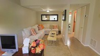 This two-floor home offers a comfortable and well-organized living environment with a welcoming aesthetic throughout. The main living area is finished in soft, neutral beige tones that create a calm and inviting atmosphere. A generously sized sectional sofa anchors the room, styled with patterned and orange accent pillows that add a pop of warmth and color. A rectangular wall mirror with a dark frame is mounted above the sofa, enhancing the sense of depth and reflecting light across the space. A sleek, light-colored ottoman serves as a central coffee table, adding a modern touch to the room.

The flooring transitions between carpet in the living area and hard surface flooring toward the hallway, providing both comfort and practicality. The open layout allows for a natural flow from the living room into a connecting hallway, which leads to additional rooms, including what appears to be a dining area with seating visible in the background. Ceiling-mounted lighting provides consistent illumination throughout. The overall design balances functionality with a cozy, lived-in charm, making this a practical and comfortable multi-level home.