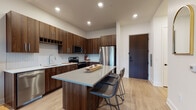 This single-floor residence showcases a thoughtfully designed contemporary interior with a cohesive aesthetic carried throughout each space. The kitchen serves as a central highlight, featuring rich walnut-toned cabinetry extending from floor to ceiling, a herringbone-patterned tile backsplash, and a full suite of stainless steel appliances including a range, microwave, and refrigerator. A generously sized center island with a light gray quartz countertop provides seating for multiple guests, making it equally functional for meal preparation and casual dining. Light hardwood flooring runs throughout the main living areas, contributing warmth and continuity. The living room is bright and inviting, benefiting from a large sliding glass door that opens to an outdoor area and allows natural light to fill the space. The bedroom offers a comfortable retreat, with a wide window fitted with blinds allowing for adjustable natural light. Neutral wall tones and plush carpeting reinforce the room's calm atmosphere. The bathroom is well-appointed with a walnut-finished vanity, white quartz countertop, an undermount sink, and a large dark-framed mirror. Recessed lighting throughout the unit ensures consistent, well-distributed illumination across all areas.