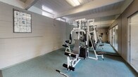 Located at 25052 Joyce St in Hayward, California, this single-story fitness center offers a well-equipped exercise space designed to support a range of workout routines. The room features a slatted white wood ceiling with overhead fluorescent lighting, complemented by high horizontal windows that allow natural light to filter in. The floor is covered in teal-toned carpet, providing a comfortable surface throughout the space. A full-length mirrored wall along one side visually expands the room and aids in monitoring form during workouts. The strength training area is anchored by a multi-station cable machine, accompanied by a curl bar and additional attachments. The cardio section includes a treadmill, elliptical trainer, and stationary spin bike, offering a variety of aerobic options. Exercise reference charts are mounted on the walls for guidance. Large sliding glass doors open directly onto a covered outdoor corridor that leads to a fenced swimming pool and surrounding patio, seamlessly connecting indoor fitness facilities with outdoor recreation. The layout is functional and straightforward, making it an accessible and practical amenity for residents of the community.
