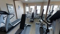 This fitness center offers a practical and well-organized workout environment designed to support a range of exercise routines. The space is housed on a single floor and benefits from abundant natural light streaming in through multiple large windows, creating a bright and energizing atmosphere. The layout accommodates both cardio and strength-training needs, featuring equipment that includes a treadmill, an elliptical or stationary bike, a multi-function cable/weight machine, and an adjustable workout bench. The flooring combines carpet and rubberized mat sections, providing a stable and comfortable surface suited to various types of training. Wall-mounted mirrors help create a sense of expanded space while allowing users to monitor their form during exercise. The neutral, light-toned walls contribute to an open and clean aesthetic. A sign near the entrance indicates that the facility is accessible during business hours, suggesting it serves as a shared amenity space. Overall, the fitness center is thoughtfully arranged to maximize usability within its footprint, offering residents or users a convenient and functional space to maintain an active lifestyle.