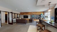 This single-floor communal space in Louisville's NuLu neighborhood combines contemporary design with warm, natural materials to create a versatile and inviting environment. The layout is open and well-organized, divided into distinct functional zones that flow seamlessly into one another.

The centerpiece of the lounge area is a striking reclaimed wood accent wall, which serves as a backdrop for a wall-mounted flat-screen television. Dark charcoal built-in shelving flanks the feature wall, providing decorative display space and a grounding visual anchor. Seating throughout includes dark leather sofas and armchairs, complemented by neutral upholstered pieces and natural wood stool ottomans, all positioned atop patterned carpet tile flooring.

A bar-style counter with a dark stone surface and pendant lighting overhead defines a separate service area, equipped with small appliances and decorative accents. Bar-height stools with leather cushions line the counter, making it suitable for casual gatherings.

A quiet dining nook features live-edge wood tables, booth-style seating, and pendant lighting, accented by a vibrant botanical moss wall installation. Large windows throughout the space bring in ample natural light and provide views of the surrounding NuLu streetscape, enhancing the overall open and welcoming ambiance.