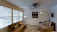 This single-floor space offers a warm and functional layout centered around a comfortable living area. The living room is finished with soft carpet flooring that adds a cozy feel underfoot, while large windows fitted with horizontal blinds allow natural light to filter in, brightening the room throughout the day. A multi-blade ceiling fan with an integrated light fixture is mounted overhead, providing both air circulation and ambient lighting. The walls are painted in a neutral, light tone that creates a clean and inviting atmosphere. A piece of abstract wall art serves as a focal point, adding a touch of personality to the space. The room comfortably accommodates plush seating, including a cushioned sofa accented with decorative pillows, making it well-suited for relaxation and everyday use. Accent lighting from floor and table lamps supplements the natural light, ensuring the space remains well-lit at any hour. The overall ambiance is casual and welcoming, with a straightforward design that prioritizes comfort and livability throughout.