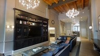 This impressive single-floor space exudes a refined, club-like atmosphere that seamlessly blends rustic and contemporary design elements. The room commands attention with its soaring ceilings, where rich, exposed wooden beams run across the upper expanse, complemented by striking modern chandeliers that cast a warm, inviting glow throughout the area. The open layout is anchored by a substantial dark wood built-in entertainment unit, complete with a large flat-screen television and flanking bookshelves stocked with books and decorative accents, lending an air of intellectual warmth to the space. Plush navy leather sofas and seating arrangements are positioned around glass-topped coffee tables, creating comfortable conversation areas that encourage relaxed gathering. Neutral walls with crisp white molding provide an elegant backdrop, balancing the darker furnishings and adding a sense of architectural refinement. Natural light filters in through large windows visible in the background, enhancing the brightness of the space. A glimpse of an adjacent area hints at additional rooms beyond, suggesting a thoughtfully connected floor plan. Overall, the space offers a sophisticated yet welcoming ambiance, ideal for socializing or unwinding in comfort.