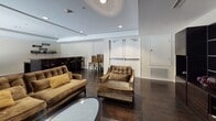 This refined single-floor space is designed with an open-plan layout that maximizes flow and functionality. Throughout the space, rich dark hardwood flooring grounds the interiors, while recessed tray ceilings with embedded lighting add architectural depth and a polished ambiance.

The main lounge area is generously proportioned, anchored by plush velvet sofas and accent chairs arranged around a sleek oval glass coffee table. A striking built-in media wall in dark wood houses a flat-screen television and display shelving, complemented by a bar counter with seating nearby — ideal for entertaining. Floor-to-ceiling dark drape curtains line the walls, adding drama and privacy, while a sliding glass door opens to an outdoor terrace, allowing natural light to filter in.

A dedicated dining nook offers an intimate setting, featuring a curved banquette with vibrant accent pillows, a round glass-top dining table, and upholstered chairs. Decorative geometric wall panels above the banquette serve as a focal point in this area, while a window with views to the exterior brings in warmth and light.

The overall ambiance of the space is upscale and cohesive, blending dark tones with warm neutrals to create an environment that is both sophisticated and inviting, suited for lounging, dining, and social gatherings alike.