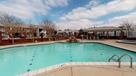 This outdoor amenity space centers around a generously sized, freeform swimming pool with clearly marked depth indicators, making it suitable for a range of swimmers. The pool's clean, tiled edges and shimmering turquoise water are complemented by a decorative rock water feature that adds a resort-like ambiance to the setting.

The surrounding pool deck is finished in a smooth concrete surface, providing ample space for lounging and socializing. A set of sling-style lounge chairs and a patio table topped with a red umbrella offer comfortable spots for relaxation in the open air. On one side of the pool, a covered pavilion with a metal roof provides shaded seating, ideal for escaping the sun while remaining close to the water.

The entire pool area is enclosed by a classic wrought iron fence with brick columns, enhancing both security and aesthetic appeal. Ornamental globe light fixtures are mounted atop the brick pillars, ensuring the space is well-lit during evening hours. Mature trees and neatly maintained shrubbery line the perimeter, offering natural privacy and a pleasant green backdrop. The surrounding building exteriors, constructed in traditional brick, frame the area and contribute to the cohesive, well-maintained character of the overall community.