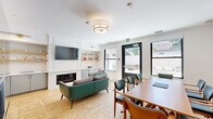 This thoughtfully designed single-floor communal space seamlessly integrates a living room, dining area, and kitchen into one cohesive, well-appointed layout. The living area anchors the space with a sleek, wall-mounted fireplace flanked by a built-in media wall featuring open wood shelving with brass-accented cabinetry below — ideal for both display and storage. A mounted flat-screen television and a mix of teal leather and tweed upholstered seating complete the lounge setting, while light-toned parquet hardwood flooring in a geometric pattern adds visual warmth and texture throughout.

Natural light flows generously through black-framed windows, offering views of an adjacent outdoor patio area accessible through a glass-paneled exit door. The dining zone accommodates a full-sized wood conference-style table surrounded by mid-century modern chairs with teal upholstery, making it a functional space for both casual gatherings and structured meetings.

The kitchen is outfitted with gray flat-front cabinetry accented with brass hardware, white quartz countertops, a glossy white subway tile backsplash, stainless steel appliances including a dishwasher and refrigerator, and open wood shelving above. The overall aesthetic is clean, contemporary, and inviting.