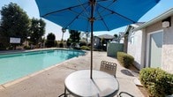 Located at 923 Alturas Road in Fallbrook, California, this outdoor pool area offers a well-maintained communal retreat designed for relaxation and leisure. The centerpiece is a generously sized swimming pool with clear, inviting water, enclosed by a secure metal fence for safety. The surrounding concrete pool deck provides ample space for poolside activities and is equipped with a shaded patio seating area featuring a round glass-top table, chairs, and a large blue market umbrella — ideal for enjoying the abundant Southern California sunshine while staying cool.

Adjacent to the pool, a landscaped lawn area provides additional outdoor space, accented by mature trees that offer natural shade and a sense of privacy. Lounge chairs are positioned throughout the grounds, inviting guests to relax and take in the open views of the surrounding hillside landscape and clear blue skies. Neatly trimmed shrubs and greenery line the pathways and building exterior, contributing to a well-kept, welcoming atmosphere.

The building exterior features a classic stucco finish with white walls, green trim, and barred windows, reflecting a practical and durable architectural style common to the region. An outdoor water fountain is conveniently mounted near the building entrance. The overall ambiance is bright, open, and conducive to outdoor enjoyment in the warm Fallbrook climate.
