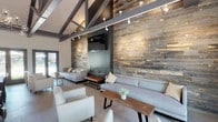 Located at 9961 Lurline Ave in the Chatsworth neighborhood of Los Angeles, this single-story space offers a thoughtfully designed interior that blends rustic and contemporary elements. The architecture is defined by dramatic vaulted ceilings with bold, dark-stained exposed beam trusses that draw the eye upward and lend the space a grand, open character.

The lounge area serves as a focal point, anchored by a striking floor-to-ceiling reclaimed wood accent wall finished in weathered grays and warm tones. A sleek, modern linear fireplace is built into this wall alongside a wall-mounted television, creating an inviting gathering spot. Comfortable neutral-toned sofas, armchairs, and a live-edge wood coffee table complete the seating arrangement.

Flowing seamlessly from the lounge is a generous dining area furnished with dark wood tables and cross-back chairs, accommodating multiple groups simultaneously. An elegant chandelier adds refinement overhead. Light wood-toned flooring runs throughout, brightening the interior alongside track lighting mounted on the beams.

Multiple sets of gray-framed French doors line one wall, flooding the space with natural light and providing direct access to an outdoor area, offering a strong indoor-outdoor connection that enhances the overall ambiance.