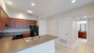 This single-floor residence offers a well-organized and functional layout designed for comfortable everyday living. The kitchen serves as a central highlight, featuring rich warm-toned wood cabinetry that provides ample storage alongside a black refrigerator and built-in appliances. A peninsula island with a neutral countertop and a double stainless steel sink anchors the space, creating a practical workspace while also serving as a natural dividing point between the kitchen and the adjacent areas. Recessed lighting illuminates the kitchen evenly, complementing the light, neutral-toned walls that lend the interior a clean and airy feel. The light-colored tile flooring extends throughout, contributing to the overall brightness of the space. Multiple white-paneled doors lead to additional rooms and closets, suggesting a thoughtful floor plan with well-distributed living areas. HVAC vents are integrated into both the walls and floor, ensuring year-round climate comfort. The overall ambiance is understated and move-in ready, with a practical design that balances form and function seamlessly across its single level.