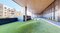 Located at 1860 Reston Row Plaza in Reston, Virginia, this contemporary outdoor terrace offers a thoughtfully designed semi-enclosed gathering space that blends architectural precision with inviting greenery. The ground is covered in vibrant artificial turf, including a gently contoured raised turf mound that adds subtle topographic interest to the level. Scattered throughout are cylindrical concrete seats that serve as minimalist, sculptural focal points, ideal for casual gatherings or relaxed individual use.

The terrace is sheltered by a warm wood-paneled ceiling with recessed lighting, providing overhead coverage while maintaining an open, airy atmosphere. Structural concrete columns punctuate the perimeter, framing views outward toward the surrounding Reston Row urban landscape and its mixed-use buildings. One side of the terrace features expansive floor-to-ceiling glazed walls with dark metal framing, offering transparency between the interior and exterior spaces. A combination of smooth stone pavers and turf creates a clear delineation between circulation paths and lounging areas.

Dark metal panel cladding lines the building's exterior facade and low perimeter barriers, contributing to the space's modern, polished aesthetic. The terrace benefits from abundant natural light and open sightlines to the sky and neighboring district, making it a versatile amenity space well suited for outdoor programming, social interaction, or quiet relaxation within a vibrant urban setting.