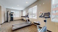 This single-floor space is divided into two distinct functional areas: a fitness room and a property management office. The fitness room is bright and welcoming, featuring large windows dressed with horizontal blinds that allow ample natural light to fill the space. The room is outfitted with a range of cardio and strength equipment, including a treadmill, stationary bike, elliptical, and a multi-function cable machine with an adjustable bench and free weights. The flooring throughout the fitness area consists of durable, patterned carpet tiles in neutral tones, and recessed ceiling lights supplement the natural light. The layout provides sufficient open floor space for comfortable movement between equipment.

Adjacent to the fitness room is the property management office, which presents a professional and organized appearance. It features a reception-style front desk with warm wood cabinetry, complemented by a secondary credenza housing office equipment. Seating for visitors consists of sleek black upholstered chairs arranged near the entrance. The office shares the same neutral wall tones and patterned carpet tiles found throughout, creating a cohesive aesthetic. A security camera is visible, and the space is accented with potted plants and artwork, contributing to an approachable, professional atmosphere.