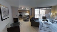 This contemporary single-floor space offers a well-designed open-concept layout that seamlessly integrates the kitchen, dining, and living areas into one cohesive environment. The kitchen is fully equipped with stainless steel appliances, including a range, microwave, and oven, complemented by warm wood-toned cabinetry and a central island on casters that provides additional prep and dining space. Track lighting overhead ensures the kitchen is well-lit and functional. The living area features a plush sofa, a glass coffee table, and a dedicated space for a television, creating a relaxed and inviting atmosphere. Light wood-toned flooring runs throughout, enhancing the bright and airy feel of the space. A standout feature is the floor-to-ceiling glass window wall adjacent to the living area, which floods the interior with natural light and provides direct access to a private balcony overlooking a vibrant urban landscape. The balcony offers an ideal spot to enjoy outdoor views of the surrounding cityscape. The overall design is modern and clean, with neutral tones and thoughtful finishes that contribute to a sophisticated yet comfortable living environment.