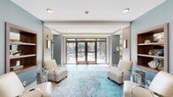 Located at 35 Folly Road Blvd in Charleston, SC, this single-floor community clubhouse offers a thoughtfully designed coastal retreat. The space is anchored by a welcoming lobby featuring light wood-look tile flooring, soft teal area rugs, and cream upholstered armchairs arranged to invite conversation, with floor-to-ceiling glass doors that open directly to an outdoor courtyard, flooding the interior with natural light.

Beyond the entry, multiple lounge areas are furnished with plush teal sectional sofas, vibrant yellow accent chairs, and colorful abstract rugs, all accented by coastal-themed artwork and pendant lighting. A dedicated media lounge with a wall-mounted television provides a relaxed entertainment option, while a shuffleboard table and a full billiards table offer additional recreation.

A standout feature is the bar area, distinguished by a striking sea-glass mosaic tile surround, quartz countertop, bar seating, pendant lights, and dual mounted televisions — creating an ideal social hub. A bright dining nook with large sliding glass windows overlooking the courtyard completes the gathering spaces. Throughout, the design maintains a cohesive coastal palette of aqua, teal, and warm neutrals, creating an inviting and resort-like atmosphere suited to the Charleston setting.