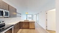 This modern single-floor residence showcases a thoughtfully designed open-concept layout that seamlessly connects the kitchen and living areas. The kitchen is a standout feature, equipped with dark-toned cabinetry, stainless steel appliances including a refrigerator, range, and built-in microwave, complemented by crisp white quartz countertops and a subway tile backsplash. A peninsula with an undermount sink and a stylish pendant light anchors the space, while open shelving adds both function and visual interest. Light wood-style flooring flows throughout the main living areas, enhancing the bright, airy atmosphere created by large windows and a full-height sliding glass door that opens to a private balcony with views of the surrounding neighborhood. A contemporary barn-style sliding door adds a modern architectural detail to the living space. The bedroom is carpeted for comfort, features a closet, and receives natural light through a tall window. The bathroom is finished with a white quartz vanity, large frameless mirror, and dark-toned cabinetry that echoes the kitchen's design language. Clean white walls and modern fixtures throughout give the entire space a cohesive, polished aesthetic.