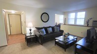 This single-floor residence offers a welcoming and well-appointed living space designed for everyday comfort. The living room serves as the central gathering area, featuring plush carpeted flooring that adds warmth underfoot. A large, cushioned sofa anchored by teal accent pillows provides a stylish and comfortable seating arrangement, complemented by a classic glass-top coffee table and a matching side table with a traditional table lamp. A round wall mirror above the sofa adds a decorative focal point and enhances the sense of depth in the room. Natural light streams in through a window, brightening the space and creating an inviting ambiance. The neutral wall tones throughout contribute to a clean, airy aesthetic that pairs well with the dark-toned furniture. A partial view into an adjacent area — likely a dining space — suggests an open, connected layout that promotes ease of movement between living areas. The hardwood flooring visible in the adjoining space provides a subtle contrast to the carpeted living room, adding visual variety to the overall design. The space strikes a balance between classic styling and functional living.