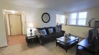 This single-floor residence offers a welcoming and well-appointed living space designed for everyday comfort. The living room serves as the central gathering area, featuring plush carpeted flooring that adds warmth underfoot. A large, cushioned sofa anchored by teal accent pillows provides a stylish and comfortable seating arrangement, complemented by a classic glass-top coffee table and a matching side table with a traditional table lamp. A round wall mirror above the sofa adds a decorative focal point and enhances the sense of depth in the room. Natural light streams in through a window, brightening the space and creating an inviting ambiance. The neutral wall tones throughout contribute to a clean, airy aesthetic that pairs well with the dark-toned furniture. A partial view into an adjacent area — likely a dining space — suggests an open, connected layout that promotes ease of movement between living areas. The hardwood flooring visible in the adjoining space provides a subtle contrast to the carpeted living room, adding visual variety to the overall design. The space strikes a balance between classic styling and functional living.