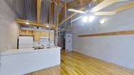 This single-floor space showcases a distinctive industrial-rustic aesthetic that sets it apart from conventional layouts. The open main area is defined by dramatically vaulted ceilings with exposed wooden beams and corrugated metal roofing, creating a sense of height and openness well beyond what the footprint might suggest. A ceiling fan with a light fixture is centrally mounted, providing both airflow and ambient lighting throughout the main living area.

Polished hardwood floors run continuously through the space, adding warmth and cohesion to the raw, textured walls, which display exposed brick and rough plaster finishes that contribute to the space's rugged, loft-like character. A bold blue entry door provides a vivid focal point, while a large window invites natural light into the living area.

The kitchen is equipped with light wood cabinetry, a white electric range, a full-size refrigerator, and a peninsula-style counter with a single-basin sink — offering practical functionality in a compact layout. Exposed ductwork and structural columns add to the industrial character of the kitchen zone.

The bathroom is clean and straightforward, featuring a single vanity with an integrated sink, a large mirror, white tile walls, and a standalone walk-in shower. Throughout the space, the interplay of raw materials and functional design delivers a utilitarian yet characterful environment.