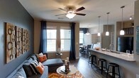 This single-floor residence showcases an open-concept layout that integrates the living, dining, and kitchen areas into one cohesive and functional space. The living area is anchored by a comfortable gray sectional sofa accented with vibrant orange and striped throw pillows, complemented by a sleek round coffee table resting on a bold chevron-patterned area rug in orange and white. A ceiling fan with dark wood-toned blades provides both style and practicality overhead. Large windows dressed with elegant navy blue drapes allow generous natural light to fill the room while adding a rich contrast to the neutral taupe walls and warm hardwood-style flooring that runs throughout.

Transitioning toward the dining area, a modest table and upholstered chairs provide a dedicated space for meals. The kitchen features a granite-topped island with bar-height seating accommodated by sleek metal stools, creating an inviting spot for casual dining or entertaining. Pendant lighting in varying styles hangs above, adding visual interest and layered illumination. The overall ambiance balances contemporary design with warm, inviting tones, resulting in a well-appointed and comfortable living environment.