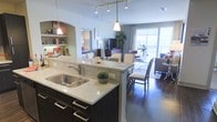This single-floor space showcases a thoughtfully designed open-concept layout that seamlessly connects the kitchen, dining, and living areas into one cohesive and functional environment. The kitchen is a standout feature, boasting dark cabinetry with sleek hardware, light stone countertops, a double basin sink, and an island with bar seating that doubles as a casual dining spot. Pendant lighting hangs above the island, adding a warm, modern touch complemented by a track lighting system overhead. Built-in shelving with an arched alcove provides additional storage and display space, adding architectural character to the kitchen area. The dining space accommodates a full table and chairs, positioned centrally between the kitchen and living room for easy flow and entertaining. The living room is open and inviting, featuring rich hardwood flooring that runs throughout the space and large sliding glass doors that flood the interior with natural light while offering access to an outdoor balcony area. The overall ambiance is contemporary and airy, with neutral tones, generous proportions, and a layout designed to maximize both comfort and functionality.