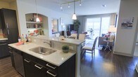 This single-floor space showcases a thoughtfully designed open-concept layout that seamlessly connects the kitchen, dining, and living areas into one cohesive and functional environment. The kitchen is a standout feature, boasting dark cabinetry with sleek hardware, light stone countertops, a double basin sink, and an island with bar seating that doubles as a casual dining spot. Pendant lighting hangs above the island, adding a warm, modern touch complemented by a track lighting system overhead. Built-in shelving with an arched alcove provides additional storage and display space, adding architectural character to the kitchen area. The dining space accommodates a full table and chairs, positioned centrally between the kitchen and living room for easy flow and entertaining. The living room is open and inviting, featuring rich hardwood flooring that runs throughout the space and large sliding glass doors that flood the interior with natural light while offering access to an outdoor balcony area. The overall ambiance is contemporary and airy, with neutral tones, generous proportions, and a layout designed to maximize both comfort and functionality.