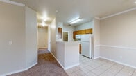 This single-floor, 2-bedroom, 1-bathroom residence offers a thoughtfully designed layout that maximizes functionality and comfort. The open-concept main living area seamlessly connects the living room, dining area, and kitchen into one cohesive space. The living room features warm neutral tones, plush carpet, crown molding, a ceiling fan with light fixture, and a window that invites natural light. The kitchen is equipped with warm wood-toned cabinetry, white appliances including a refrigerator and range, and a peninsula breakfast bar that doubles as a convenient divider between the kitchen and living spaces. Light tile flooring runs throughout the kitchen and entry areas. A dedicated pantry provides additional storage adjacent to the kitchen. Both bedrooms are carpeted and feature neutral walls, windows with blinds for natural light and privacy, and access to closets — including a spacious walk-in closet with built-in shelving and hanging rods. The bathroom includes a full tub-shower combination with tiled surround, toilet, and a vanity with a countertop. A dedicated laundry area with washer and dryer hookups adds everyday convenience. A hallway connects the main living areas to the bedrooms, utility room, and additional closet storage, ensuring a practical and organized flow throughout the home.