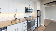 This thoughtfully designed single-floor space showcases a compelling blend of industrial and contemporary aesthetics. The open-concept living area is immediately striking, featuring soaring exposed concrete ceilings with visible structural steel beams that lend a raw, urban character to the space. Warm hardwood-style flooring flows seamlessly throughout, complementing the neutral color palette of the walls.

Floor-to-ceiling windows line one wall of the living area, flooding the space with abundant natural light and offering expansive views of the surrounding cityscape. Exposed ductwork and concrete columns further enhance the loft-like, industrial ambiance.

The kitchen is well-appointed with crisp white shaker-style cabinetry extending to the ceiling, maximizing storage. Stainless steel appliances — including a range, over-the-range microwave, and refrigerator — pair with a classic white subway tile backsplash and dark countertops. Matte black hardware and a black-finish faucet provide sleek, modern accents throughout.

The full bathroom is clean and contemporary, featuring a tiled tub-and-shower combo, a white vanity with an integrated sink, and an illuminated LED mirror. Matte black fixtures are consistent with the design language found throughout the unit. A separate water closet provides additional privacy and convenience.