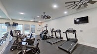 This single-level fitness center offers a comprehensive workout environment designed to accommodate a wide range of exercise routines. The space is thoughtfully laid out to maximize functionality, with distinct zones for cardio training, strength work, and functional fitness. The cardio area is outfitted with multiple treadmills, a stair climber, and stationary cycling bikes, providing ample options for aerobic exercise. A wall-mounted flat-screen television is positioned for viewing during workouts.

The strength training section features a full rack of dumbbells spanning a wide weight range, a weight bench, and a cable machine, supporting both targeted and compound exercises. Medicine balls and stability balls are neatly stored on a dedicated rack, adding versatility for functional training. Large floor-to-ceiling mirrors line one wall, visually expanding the space and allowing users to monitor their form throughout their workout.

The interior finishes are clean and modern, with crisp white walls, recessed lighting, and dark rubber flooring that provides durability and traction. Distinctive windmill-style ceiling fans contribute both airflow and a stylish aesthetic detail. Natural light enters through windows and glass doors that connect to an adjacent corridor, keeping the environment bright and inviting.