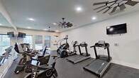 This single-level fitness center offers a comprehensive workout environment designed to accommodate a wide range of exercise routines. The space is thoughtfully laid out to maximize functionality, with distinct zones for cardio training, strength work, and functional fitness. The cardio area is outfitted with multiple treadmills, a stair climber, and stationary cycling bikes, providing ample options for aerobic exercise. A wall-mounted flat-screen television is positioned for viewing during workouts.

The strength training section features a full rack of dumbbells spanning a wide weight range, a weight bench, and a cable machine, supporting both targeted and compound exercises. Medicine balls and stability balls are neatly stored on a dedicated rack, adding versatility for functional training. Large floor-to-ceiling mirrors line one wall, visually expanding the space and allowing users to monitor their form throughout their workout.

The interior finishes are clean and modern, with crisp white walls, recessed lighting, and dark rubber flooring that provides durability and traction. Distinctive windmill-style ceiling fans contribute both airflow and a stylish aesthetic detail. Natural light enters through windows and glass doors that connect to an adjacent corridor, keeping the environment bright and inviting.