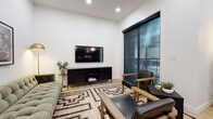 This single-floor space showcases a thoughtfully designed contemporary interior with a cohesive dark-and-light aesthetic carried throughout. The open-concept kitchen serves as a centerpiece, featuring matte black cabinetry, a large waterfall-edge island with seating for multiple guests, stainless steel appliances, a hexagonal tile backsplash, and globe pendant lighting. Large windows frame urban streetscape views, flooding the space with natural light. Light-toned hardwood flooring flows seamlessly through the kitchen and into the adjoining living area.

The living room is warmly appointed with a tufted sage green sofa, leather accent chairs, and a bold geometric area rug. A wall-mounted flat-screen TV sits above a sleek black media console, while framed black-and-white photography and a brass floor lamp add curated character to the space.

The bedroom continues the modern aesthetic with a floor-to-ceiling window offering city views, crisp white bedding accented by a chunky knit throw, and paired framed botanical prints above the headboard.

The bathroom maintains the dark-toned design language with slate tile flooring, a matte black vanity, and a tub-shower combination, creating a clean and functional space.