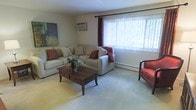 This single-floor space features a welcoming living room designed for comfort and functionality. The room is finished with wall-to-wall carpeting in a neutral tone, creating a warm and cohesive foundation throughout. A large picture window dressed with sheer vertical blinds and accent drapes allows generous natural light to fill the space while offering views of the surrounding greenery outdoors.

The living room is generously sized, accommodating a sectional sofa and additional seating with ease, centered around a dark wood coffee table that adds a grounding contrast to the light color palette. Accent pillows and a throw blanket introduce warm red tones, providing visual interest against the neutral furnishings.

Climate comfort is well-supported, with a wall-mounted air conditioning unit positioned above the sofa and a baseboard heating unit running along the exterior wall beneath the window, ensuring year-round temperature control. The walls are finished in a clean, neutral tone, complemented by a bold abstract artwork that adds a decorative focal point. Overall, the space presents a practical and comfortable living environment with a relaxed, residential ambiance.