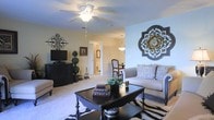 This single-floor residence offers a thoughtfully designed living space that balances comfort and style. The living room serves as the heart of the home, featuring plush wall-to-wall carpeting, light-toned walls, and a ceiling fan with integrated lighting that ensures year-round comfort. A bold zebra-print area rug anchors the seating arrangement, which centers around a dark wood coffee table adorned with decorative books and greenery. The sofa is dressed with patterned throw pillows, adding texture and warmth to the room. Striking wall décor, including an ornate medallion piece and an iron cross accent, lend the space a sophisticated, curated character. A media console with a flat-screen television is positioned along one wall, complemented by decorative topiaries that add a touch of greenery. The open layout connects seamlessly to what appears to be a dining area, visible in the background and illuminated by a pendant light fixture. A glimpse through an interior doorway reveals additional rooms, including what appears to be a bathroom with a mirror, suggesting a well-connected and functional floor plan throughout. The overall ambiance is warm, elegant, and inviting.