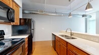 Located at 2828 Euclid Ave in Cleveland, Ohio, this one-bedroom, one-bathroom residence offers a distinctive urban-industrial aesthetic throughout. The open-concept kitchen and living area forms the heart of the unit, featuring warm bamboo-style hardwood flooring, exposed silver ductwork running along the ceiling, and pendant lighting that adds a modern industrial character. The kitchen is well-appointed with rich wood cabinetry, light countertops, a double stainless steel sink, and black appliances including a refrigerator, range, microwave, and dishwasher. A breakfast bar separates the kitchen from the living area, which benefits from natural light provided by large windows with vertical blinds offering views of the surrounding urban streetscape. A narrow hallway with the same exposed ductwork detail connects the main living space to the private areas of the unit. The bedroom features soft carpeting, a ceiling fan, and a built-in overhead shelf, creating a comfortable and functional retreat. The bathroom includes a stand-up shower, a wood-cabinet vanity, and durable hardwood-style flooring, maintaining a clean and consistent design language throughout the unit. The overall layout is efficiently organized, making excellent use of the available footprint.