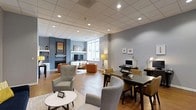 Located on Steinbeck Drive in Raleigh, North Carolina, this single-floor community space offers a thoughtfully designed environment that blends comfort with functionality. The open-concept layout seamlessly connects a relaxed lounge area with a dedicated co-working zone, making it well-suited for both socializing and focused work.

The lounge area features a built-in shelving unit with a fireplace surround finished in a deep blue-gray tone, serving as a striking focal point. A wall-mounted television is framed by accent lighting and curated décor, while warm hardwood flooring throughout adds character and continuity. Plush armchairs in neutral and earthy tones — including rust orange and soft gray — are arranged around a marble-top coffee table, creating an inviting gathering spot.

Large windows allow natural light to fill the space, complemented by stylish floor lamps that provide a warm ambient glow. The co-working area includes sleek dark-finished desks equipped with desktop computers, task lighting, and ergonomic seating in mustard yellow and gray, encouraging productivity.

The suspended tile ceiling with recessed lighting ensures even, comfortable illumination throughout. The overall ambiance is modern yet approachable, with a carefully curated color palette of cool grays, warm wood tones, and bold accent hues that give the space a polished, welcoming character.