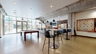 Located at 2838 Fremont Ave S in Minneapolis, MN, this contemporary club room is a well-appointed communal space designed for socializing and recreation. The open, single-floor layout is anchored by polished concrete floors that reflect an abundance of natural light pouring in through expansive floor-to-ceiling windows overlooking the surrounding greenery.

At the heart of the space sits a full kitchen, featuring dark wood cabinetry, light stone countertops, a warm butcher block bar counter, stainless steel appliances including a refrigerator and dishwasher, and a wall-mounted television — making it well-suited for hosting gatherings. A row of bar-height seating lines the kitchen counter, complemented by a freestanding high-top table and additional bar stools in the main lounge area.

Exposed ductwork and industrial-style pendant lighting add a modern, urban character to the ceiling, while a striking large-scale mural on one wall brings bold artistic energy to the room. A decorative textured wall panel frames a built-in media center and lounge bar area, creating a distinct entertainment zone. A full-size billiards table serves as a central recreational focal point. The space also provides access to an adjacent outdoor patio area, further extending its entertaining potential.