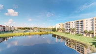 This single-floor property is situated within a well-established residential community that offers a range of notable outdoor amenities and scenic surroundings. The exterior views reveal a picturesque, calm pond that serves as a central focal point of the community, its still waters reflecting the multi-story residential buildings and swaying palm trees that line the grounds. The landscape is lush and meticulously maintained, with broad stretches of green lawn extending alongside the water's edge.

Beyond the pond, the community features tennis courts, providing recreational opportunities directly on-site. The surrounding buildings present a warm, neutral-toned facade typical of South Florida architecture, with balconies that overlook the serene water and greenery. Mature palm trees are distributed throughout the grounds, lending a tropical character to the overall setting.

The community's layout promotes an open, park-like atmosphere, with sightlines extending across the water toward the courts and neighboring structures. The combination of the reflective pond, verdant landscaping, and recreational facilities creates a peaceful and resort-like ambiance. The property benefits greatly from its position within this community, where natural beauty and outdoor living are central to the daily experience.