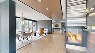 Located at 590 Park St in Saint Paul, Minnesota, this sophisticated single-floor amenity space offers a generous, open layout designed for socializing and relaxation. The interior is defined by a striking double-sided fireplace serving as the architectural centerpiece, clad in dark stone tile with built-in shelving and a bold striped surround that draws the eye from every angle. Warm wood-plank flooring flows throughout, complemented by a combination of wood-paneled ceiling sections and crisp white finishes that create a balanced, modern aesthetic. The living area features curated contemporary seating, including leather accent chairs and a textured area rug, anchored beside a textured stone feature wall that adds depth and dimension. Large floor-to-ceiling windows flood the lounge with natural light while framing views of the surrounding neighborhood. A dedicated game room area accommodates a full-size pool table beneath stylish pendant lighting, positioned adjacent to a sleek wet bar with quartz countertops, bar seating, and a beverage cooler. A slatted wood room divider provides architectural interest near a utility sink area finished with geometric tile. The space also includes a snack and retail display area, a wall-mounted TV, and ample room for gatherings, making it a well-appointed communal destination.