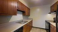 This single-floor space offers a straightforward and functional layout across its key areas. The main room features wall-to-wall carpeting in a neutral tone, complemented by light beige walls and a clean white ceiling. A ceiling-mounted HVAC vent ensures climate control, and natural light filters into the room, casting soft patches of sunlight across the floor. Dark baseboards provide a subtle contrast against the light-colored walls, adding a degree of visual definition to the space.

The kitchen is equipped with classic medium-toned wood cabinetry, offering ample upper and lower storage. The countertops are finished in a light gray laminate surface, providing practical workspace. A stainless steel double-basin sink is centrally positioned along the counter, fitted with a standard faucet. The kitchen also includes a dishwasher and features tile flooring in a neutral earthy tone. An electrical panel access door is visible on one wall, keeping utility components conveniently contained. The overall ambiance of the space is simple, clean, and ready to be personalized.