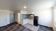 This single-floor unit offers a thoughtfully organized layout that combines the living and kitchen areas into one open space, maximizing functionality. The kitchen is positioned along one wall in an L-shaped configuration and features white shaker-style cabinetry with granite-look countertops, an undermount sink, a smooth electric cooktop, an overhead range hood, and a full-size black refrigerator. A distinctive textured metallic backsplash adds a contemporary accent to the kitchen zone. The transition between the kitchen and living area is defined by a shift from light wood-look luxury vinyl plank flooring to a patterned, multi-tone carpet that covers the main living space, adding warmth and visual interest. The living area is generously proportioned with neutral white walls and a window fitted with a wall-unit HVAC system, providing natural light and climate control. A dedicated entryway near the unit door includes a closet for added storage. The bathroom is cleanly finished with a walk-in shower featuring a glass panel, a pedestal-style white vanity with under-cabinet storage, a toilet, and a backlit LED vanity mirror that provides both functional and ambient lighting. Chrome fixtures and wood-look flooring complete the bathroom's modern, clean aesthetic.