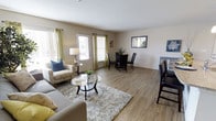 This single-story space features an open-plan layout that seamlessly connects the living and dining areas, creating a cohesive and functional environment. The light-toned, wood-look flooring extends throughout, lending warmth and visual continuity to the entire space.

The living area is well-proportioned, accommodating a comfortable sofa and an accent armchair accented with a blue throw pillow. A round glass-top coffee table sits atop a plush, textured area rug, adding a layered, inviting feel underfoot. Natural light flows generously through multiple windows dressed with yellow-green grommet curtains, brightening the neutral beige walls and contributing to an airy atmosphere.

The dining area is positioned adjacent to the living space and features a glass-top dining table surrounded by dark-framed chairs, providing an elegant yet understated dining setting. A framed mirror on the wall enhances the sense of openness. Additional bar-height seating is visible along one side, offering versatile dining options.

Overall, the space presents a clean, contemporary aesthetic with a warm, welcoming ambiance, well-suited for everyday living and casual entertaining.
