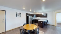 This single-floor space offers a clean, modern aesthetic with an open-concept layout that seamlessly connects the kitchen and dining areas. The kitchen is a standout feature, boasting rich dark espresso cabinetry that extends to the ceiling, complemented by light granite-style countertops that provide a striking contrast. Stainless steel appliances, including a refrigerator and range, lend a polished, contemporary feel, while a peninsula with bar seating adds both functionality and a natural gathering point. A pendant light fixture anchors the kitchen space with a warm, focused glow, and recessed lighting throughout ensures the entire area is well-lit. The dining area comfortably accommodates a round wooden table with seating for four, creating an inviting space for meals and conversation. Light wood-style laminate flooring runs consistently across the open floor plan, enhancing the sense of continuity and space. The neutral white walls serve as a clean backdrop, allowing the dark cabinetry and warm wood tones to take center stage. A framed piece of wall art adds a subtle decorative touch. Overall, the space presents a well-appointed, move-in-ready environment with a cohesive and contemporary design sensibility.