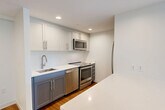This well-appointed 2-bedroom, 1-bathroom residence in Cambridge, MA offers a thoughtfully designed layout that maximizes every inch of its compact footprint. The open-concept living and dining area is anchored by warm hardwood flooring and flanked by large, floor-to-ceiling windows that flood the space with natural light and frame sweeping views of the Cambridge and Boston skyline, including treetops and surrounding urban architecture.

The modern kitchen is a standout feature, outfitted with two-tone cabinetry — white upper cabinets paired with grey lower cabinets — complemented by crisp white quartz countertops and a full suite of stainless steel appliances, including a range, dishwasher, microwave, and refrigerator. A central kitchen island provides additional prep and storage space, seamlessly connecting the kitchen to the living area.

Both bedrooms are carpeted and bright, benefiting from generous windows with city views and ample closet space. The full bathroom is clean and contemporary, featuring white cabinetry, a quartz vanity, and a tub-shower combination. Additional conveniences include in-unit laundry and a hallway connecting all rooms efficiently. The overall design aesthetic is clean, modern, and move-in ready.