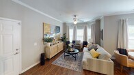 Located at 9325 Blue House Rd in North Charleston, SC, this well-appointed single-floor residence offers a thoughtfully designed layout with multiple bedrooms, two full bathrooms, a welcoming living area, and a fully equipped kitchen. The living room is a standout space, anchored by a distinctive bay window arrangement that floods the room with natural light. Rich hardwood-style flooring, crown molding, and a ceiling fan contribute to the room's warm, inviting ambiance. The kitchen features dark cabinetry, granite countertops, black appliances including a refrigerator, range, and microwave, along with track lighting and a pantry with double doors. Each bedroom is carpeted and equipped with a ceiling fan, offering a comfortable retreat. The primary bedroom features a tufted headboard, ample closet space, and natural light through slatted blinds. A secondary bedroom mirrors this comfort with neutral tones and tasteful décor. Both bathrooms are finished with dark wood vanities, granite countertops, and tub-shower combinations with wood-style flooring. A connecting hallway ties the layout together efficiently, making this residence a functional and visually cohesive living space.