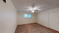 This single-floor home offers a well-organized layout with multiple rooms connected by open doorways, creating a cohesive and functional flow throughout the space. The living areas are finished with warm brown carpet and neutral cream-toned walls, lending a comfortable and understated ambiance. Crown molding and textured ceilings add a classic architectural detail to several rooms, while ceiling fans with integrated light fixtures are present throughout, ensuring both comfort and consistent illumination.

The bedrooms are generously sized, each featuring plush carpeting, multiple windows dressed with horizontal blinds, and ceiling fans that fill the rooms with natural light from the surrounding greenery visible outside. The open floor plan doll's-eye view reveals a thoughtfully arranged layout with multiple interconnected rooms and louvered closet doors providing convenient storage.

The kitchen is equipped with warm honey-toned cabinetry, a gas range, built-in microwave, full-size refrigerator, and a double-basin sink — all finished in white. Tile flooring and a window overlooking the exterior make the kitchen a bright and practical workspace.

The bathroom features a wide vanity with warm wood cabinetry, a large mirror, tile flooring, and direct access to an adjacent carpeted room, making it a convenient and well-appointed space. Overall, the home presents a clean, move-in-ready environment with consistent finishes throughout.