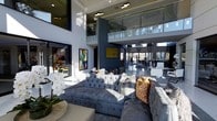 This striking two-story space showcases a bold contemporary design defined by its dramatic double-height ceilings and an open-plan layout that creates an expansive, airy atmosphere. The main living area draws immediate attention with a large, tufted velvet sectional sofa in steel blue, accented with textured fur and leopard-print cushions, reflecting a confident, design-forward aesthetic.

Large floor-to-ceiling windows and glazed walls flood the interior with natural light, highlighting the clean lines and dark navy accent walls that contrast elegantly with bright white surfaces. A glass-paneled mezzanine level overlooks the main living space below, connected by a modern railing system that preserves sightlines and reinforces the sense of vertical volume.

A sculptural sputnik-style chandelier hangs overhead, adding a focal point to the upper volume. Framed artwork adorns the walls, contributing color and artistic character. The adjoining areas, visible through the open layout, include what appears to be a bar or reception-style counter, further enhancing the sophisticated, upscale ambiance of this well-appointed two-level space.