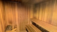 This single-level sauna is constructed entirely from natural cedar wood, creating a warm, enveloping atmosphere characteristic of traditional Scandinavian-style heat rooms. The walls, ceiling, and flooring are all finished in vertical and horizontal cedar planking, which not only contributes to the room's rich, golden-amber aesthetic but also serves a functional purpose — cedar is naturally resistant to moisture and helps regulate humidity within the space.

The interior features tiered wooden benches arranged along the walls, allowing users to choose their preferred heat level, as temperatures rise toward the ceiling. The lower and upper bench configurations provide flexibility for sitting or reclining during sessions. A traditional sauna heater is positioned in the corner, serving as the focal point of the room and the primary source of heat. The heater's stone-filled basket is designed to accept water, allowing users to generate steam and control the humidity level to their preference.

The compact dimensions of the sauna create an intimate, cocoon-like environment ideal for relaxation and therapeutic heat exposure. Subdued, warm lighting enhances the overall ambiance, reinforcing the sense of calm and privacy within the space.