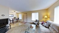 This single-floor residence offers a comfortable and functional layout centered around an open-concept living and dining area. The living room is generously sized, featuring plush carpeting and a large window dressed with vertical blinds that allows ample natural light to fill the space. A glass-topped coffee table and a sofa with accent pillows contribute to the room's welcoming atmosphere, while a cushioned accent chair and a floor lamp add practical style to the corner. A ceiling fan with an integrated light fixture is positioned overhead, providing both comfort and additional illumination. Adjacent to the living room, the dining area transitions to a tile floor and accommodates a small dining table with chairs, positioned near another large window that further brightens the space. The two areas are seamlessly connected, making the layout ideal for everyday living and casual entertaining. Warm yellow-toned walls unify the open space, creating a cozy, inviting ambiance throughout. A paneled entry door is centrally located, offering convenient access. The textured ceiling adds a classic residential character to the overall design of this practical and well-appointed home.