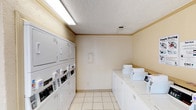This shared laundry facility is a well-equipped, single-floor space designed to serve a residential community. The room is finished with light ceramic tile flooring and warm beige walls accented by a simple crown molding detail, creating a clean and functional environment. A textured ceiling with recessed ventilation and overhead fluorescent lighting ensures the space is well-lit throughout.

The facility houses multiple commercial-grade top-loading washing machines arranged along one wall, each equipped with individual coin-operated or card-reader payment systems. Along the opposite wall, stacked dryer units maximize the use of vertical space, accommodating a high volume of laundry needs efficiently. Instructional signage from the service provider is posted on the walls for user guidance.

A small fold-down countertop is available for sorting or folding laundry, and a cork bulletin board is mounted near the entrance for community notices. A glass-paned exterior door with an adjacent large window allows natural light to filter into the room while providing a view of and access to an outdoor area. The overall ambiance is practical and straightforward, prioritizing utility and ease of use for residents.