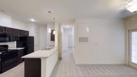 This single-floor residential space is designed with a clean, modern aesthetic and an open-concept layout that promotes a sense of spaciousness and flow. The kitchen serves as a central feature, anchored by a peninsula with light granite-style countertops and dark cabinetry, creating a striking contrast. Pendant lighting hangs above the peninsula, adding a contemporary touch and providing focused task lighting. The kitchen is equipped with a stainless steel sink and includes a full-size refrigerator. Soft, neutral-toned carpeting covers the main living area, contributing to a warm and comfortable atmosphere. Crisp white walls throughout the space enhance the brightness and maintain a cohesive, minimalist look. A hallway leads from the main living area toward additional rooms, with recessed lighting overhead guiding the way. Doors along the corridor suggest access to private spaces such as bedrooms or a bathroom. A wall-mounted thermostat and a ceiling ventilation unit reflect the practical, well-equipped nature of the unit. The overall ambiance is bright, clean, and move-in ready, making it a functional and stylish living environment.