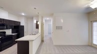 This single-floor residential space is designed with a clean, modern aesthetic and an open-concept layout that promotes a sense of spaciousness and flow. The kitchen serves as a central feature, anchored by a peninsula with light granite-style countertops and dark cabinetry, creating a striking contrast. Pendant lighting hangs above the peninsula, adding a contemporary touch and providing focused task lighting. The kitchen is equipped with a stainless steel sink and includes a full-size refrigerator. Soft, neutral-toned carpeting covers the main living area, contributing to a warm and comfortable atmosphere. Crisp white walls throughout the space enhance the brightness and maintain a cohesive, minimalist look. A hallway leads from the main living area toward additional rooms, with recessed lighting overhead guiding the way. Doors along the corridor suggest access to private spaces such as bedrooms or a bathroom. A wall-mounted thermostat and a ceiling ventilation unit reflect the practical, well-equipped nature of the unit. The overall ambiance is bright, clean, and move-in ready, making it a functional and stylish living environment.